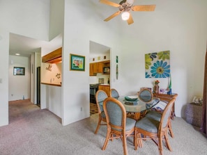 A dining area with a round table and wicker chairs, a ceiling fan, and a kitchen in the background.