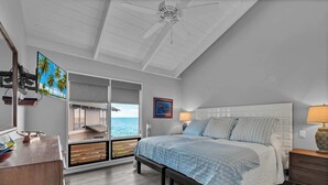 Ein Schlafzimmer mit Bett, Nachttischen, Deckenventilator, Fenster mit Meerblick und einem an der Wand befestigten Fernseher.