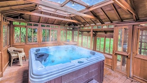 Un jacuzzi en una terraza de madera con paredes de vidrio y vistas a la vegetación.