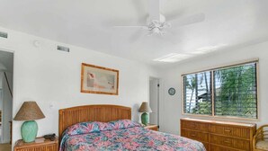 Un dormitorio con una cama grande, un ventilador de techo, una ventana con persianas, un aparador de madera y una mesita de noche con lámpara.