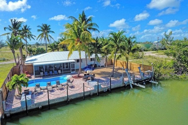 A house with a pool, surrounded by palm trees, and a dock extending into a body of water.