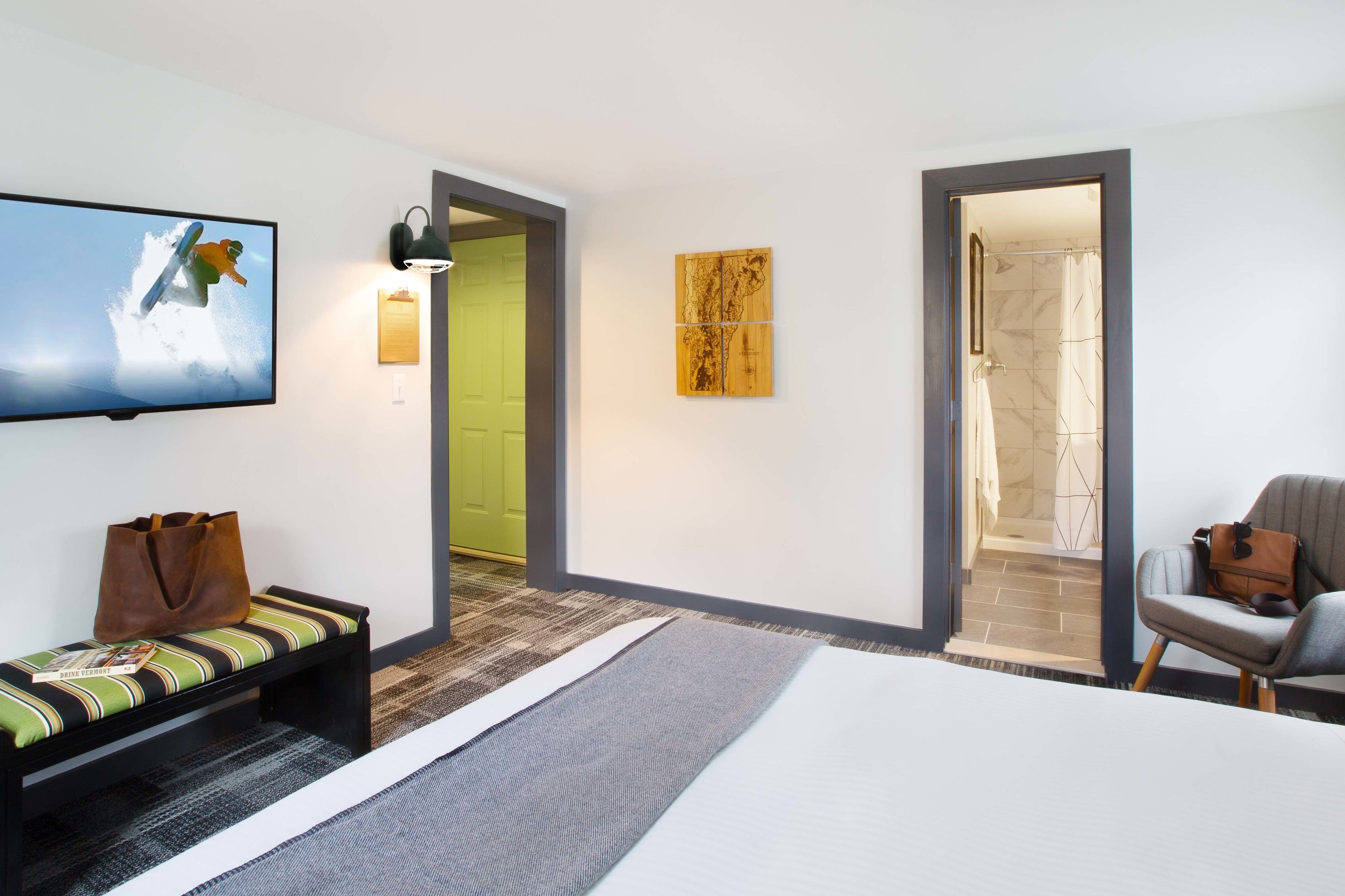 A hotel room with a bed, a bench with a bag, a TV, a wall art piece, and a shower area.