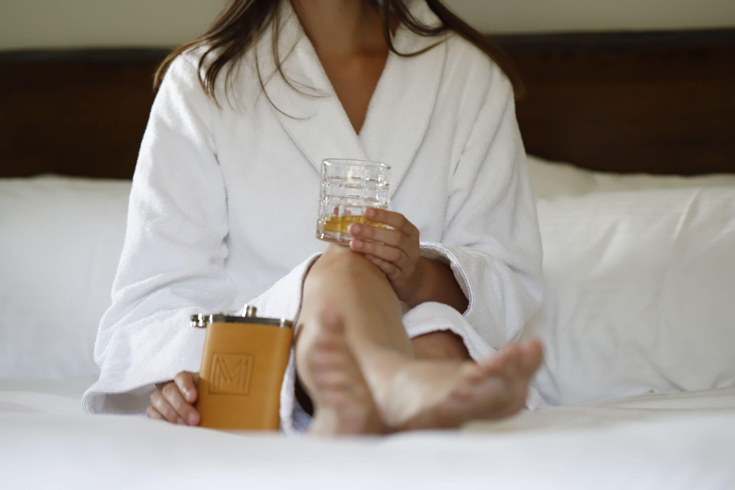 A person sitting on a bed in a white robe, holding a glass of amber liquid and a small brown object.