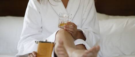 A person sitting on a bed in a white robe, holding a glass of amber liquid and a small brown object.