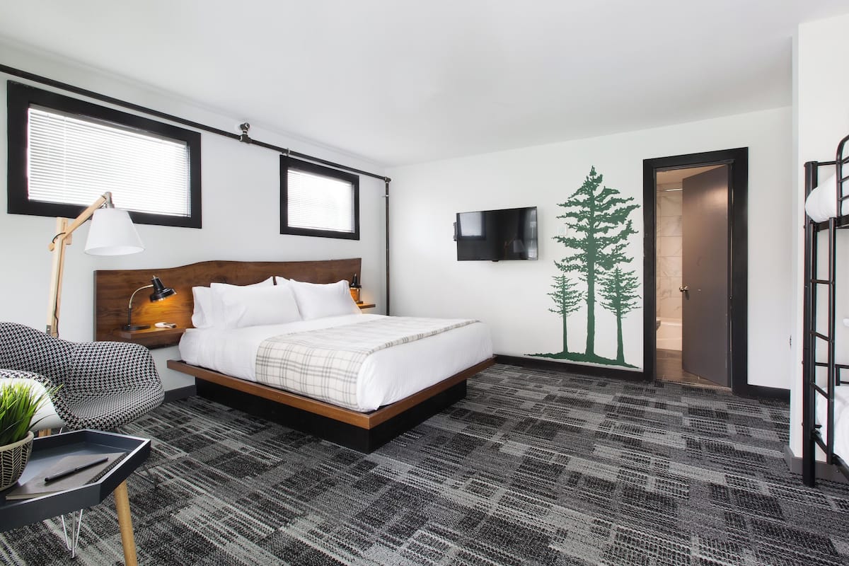 A hotel room with a bed, a desk with a lamp, a TV, a plant, and a bunk bed.