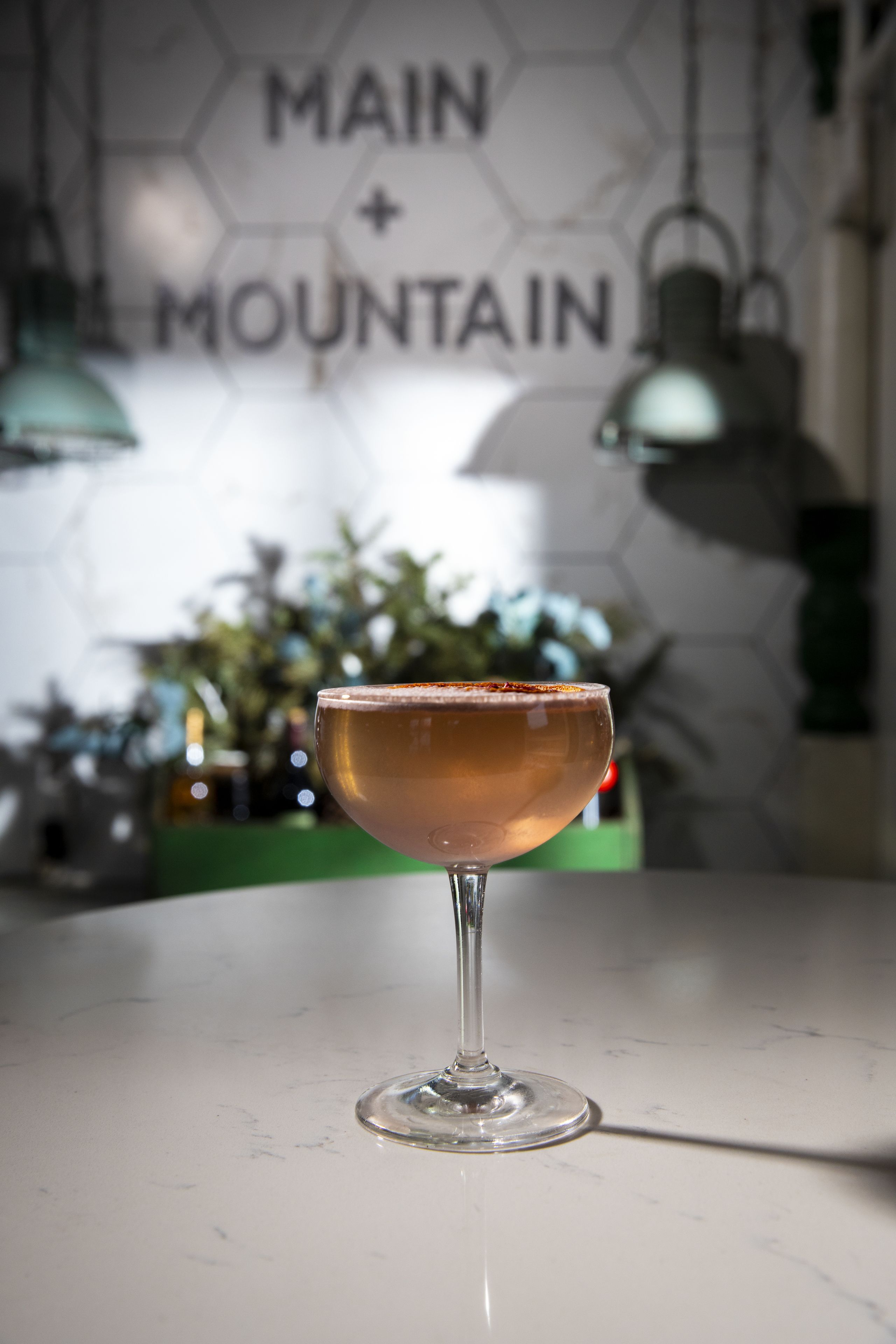 A cocktail glass on a marble countertop with a hexagonal wall pattern and a sign reading "MAIN + MOUNTAIN".