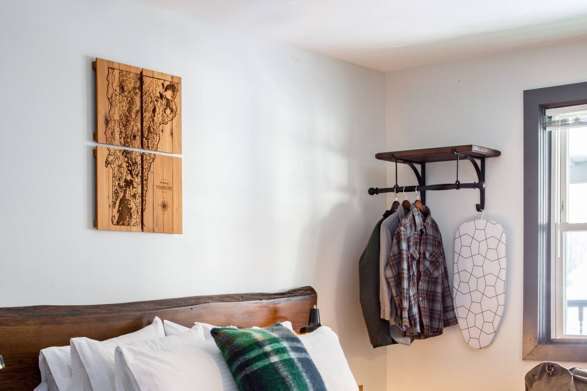 A neatly made bed with white linens and a green and blue plaid pillow, a wooden headboard, a wall-mounted coat rack with clothes, and a map artwork.
