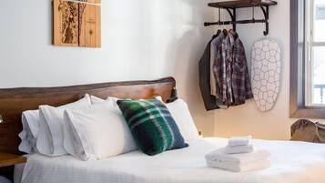 Un lit bien fait, avec du linge de lit blanc et un coussin à carreaux verts et bleus, une tête de lit en bois, un porte-manteau mural avec des vêtements et une œuvre d’art représentant une carte.