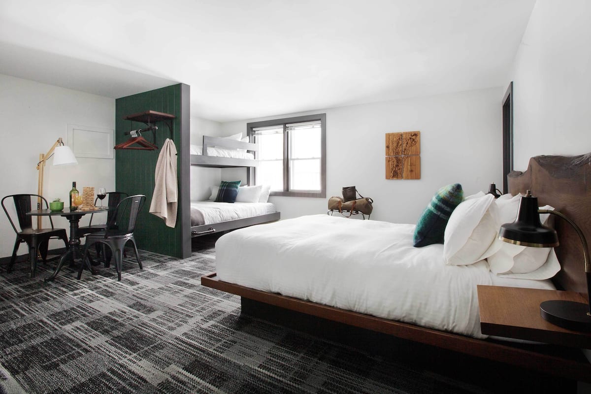 A modern hotel room with a large bed, a desk with a chair, a small table, a window, and a wall-mounted TV.