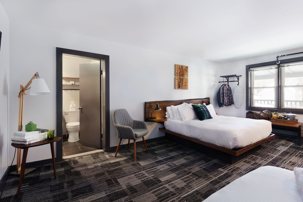 A modern hotel room with a large bed, a desk with a lamp, a chair, a wardrobe, and a window with a view.