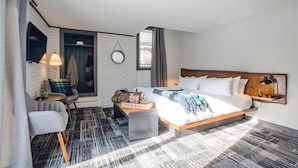 A modern hotel room with a bed, a seating area with a chair and a small table, a TV mounted on the wall, and a window with curtains. - Main and Mountain Bar & Motel (Ludlow)