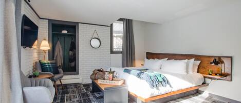 Une chambre d’hôtel moderne comprenant un lit, un coin salon avec un fauteuil et une petite table, un téléviseur fixé au mur et une fenêtre avec des rideaux.