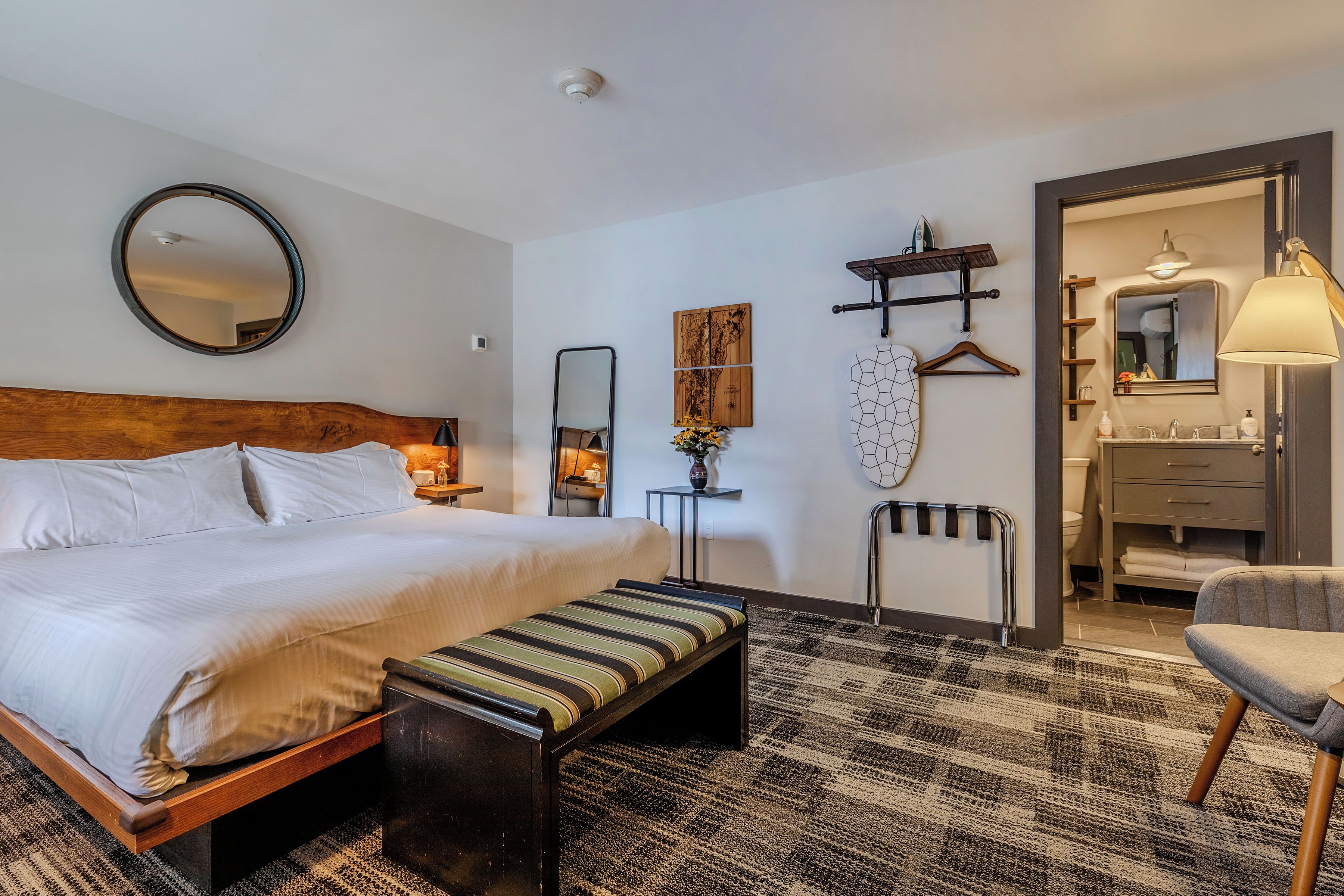 A hotel room with a bed, a bench, a mirror, a map, a towel rack, a hanging lamp, and a bathroom with a sink and mirror.