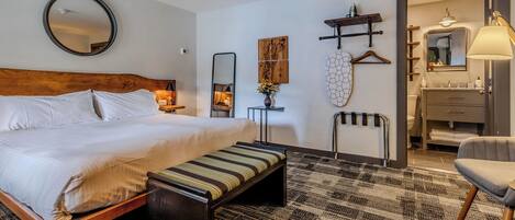 A hotel room with a bed, a bench, a mirror, a map, a towel rack, a hanging lamp, and a bathroom with a sink and mirror.