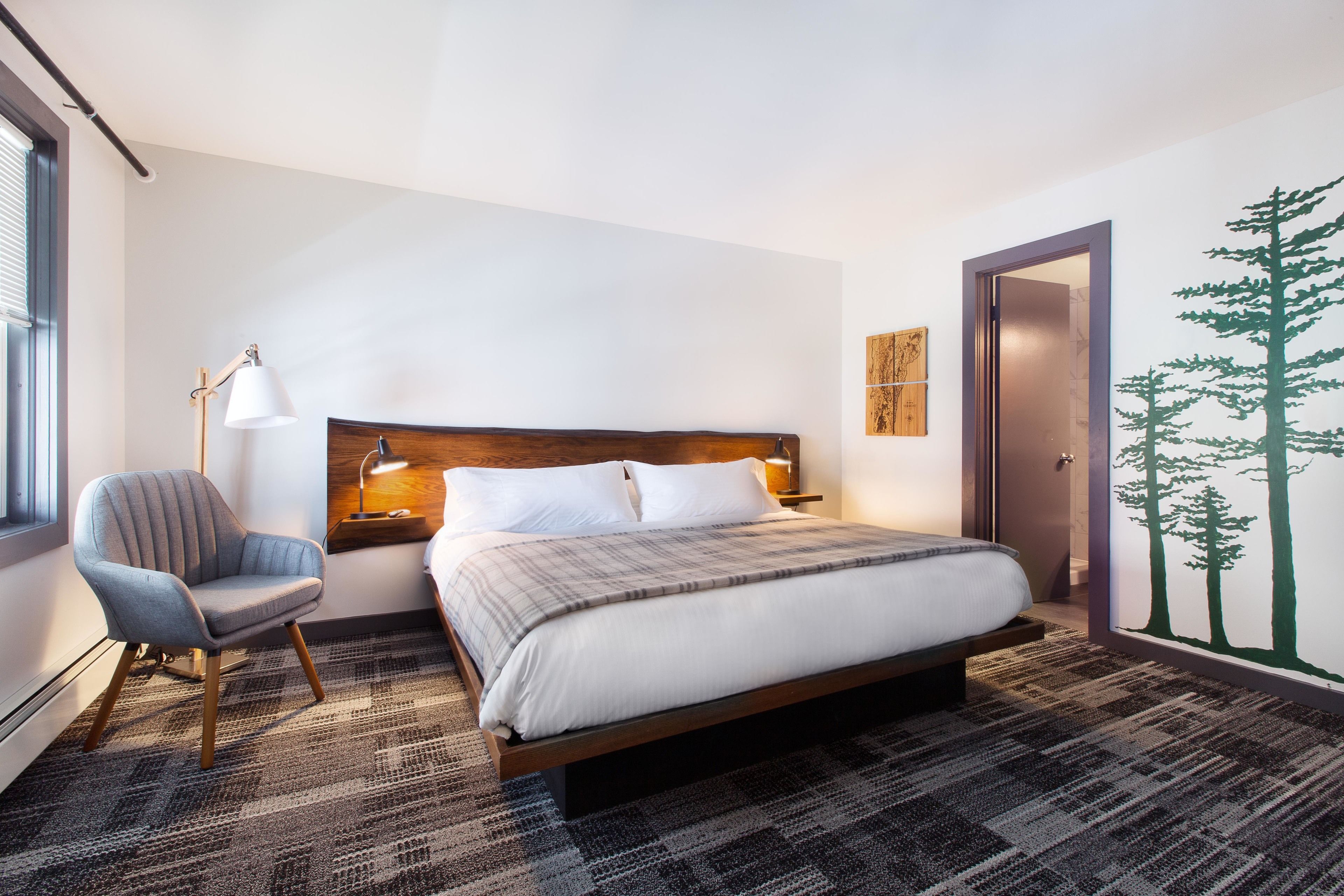 Una habitación de hotel moderna con una cama grande, una silla, una mesita de noche con lámpara y un mural en la pared.