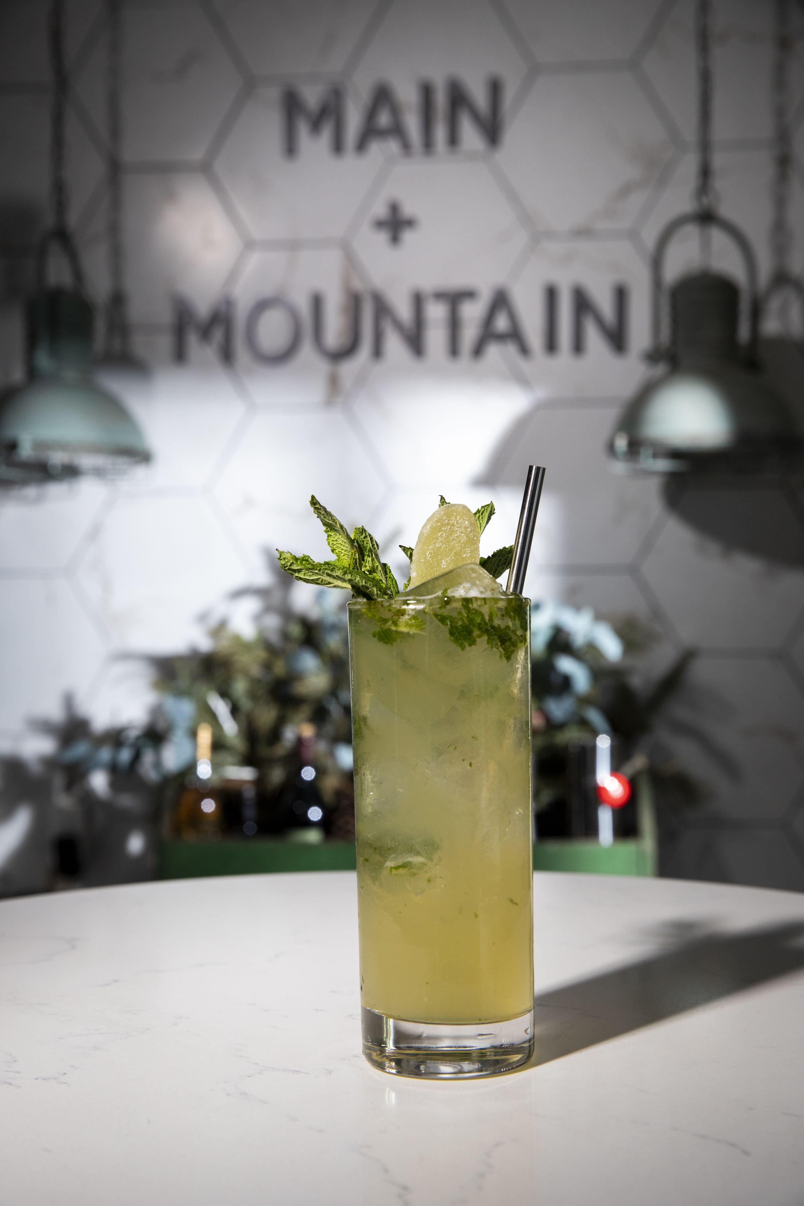 A tall glass of a light-colored beverage with mint leaves and a lemon slice, placed on a white table with a hexagonal patterned wall in the background.