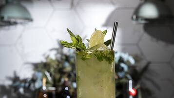 A tall glass of a light-colored beverage with mint leaves and a lemon slice, placed on a white table with a hexagonal patterned wall in the background.