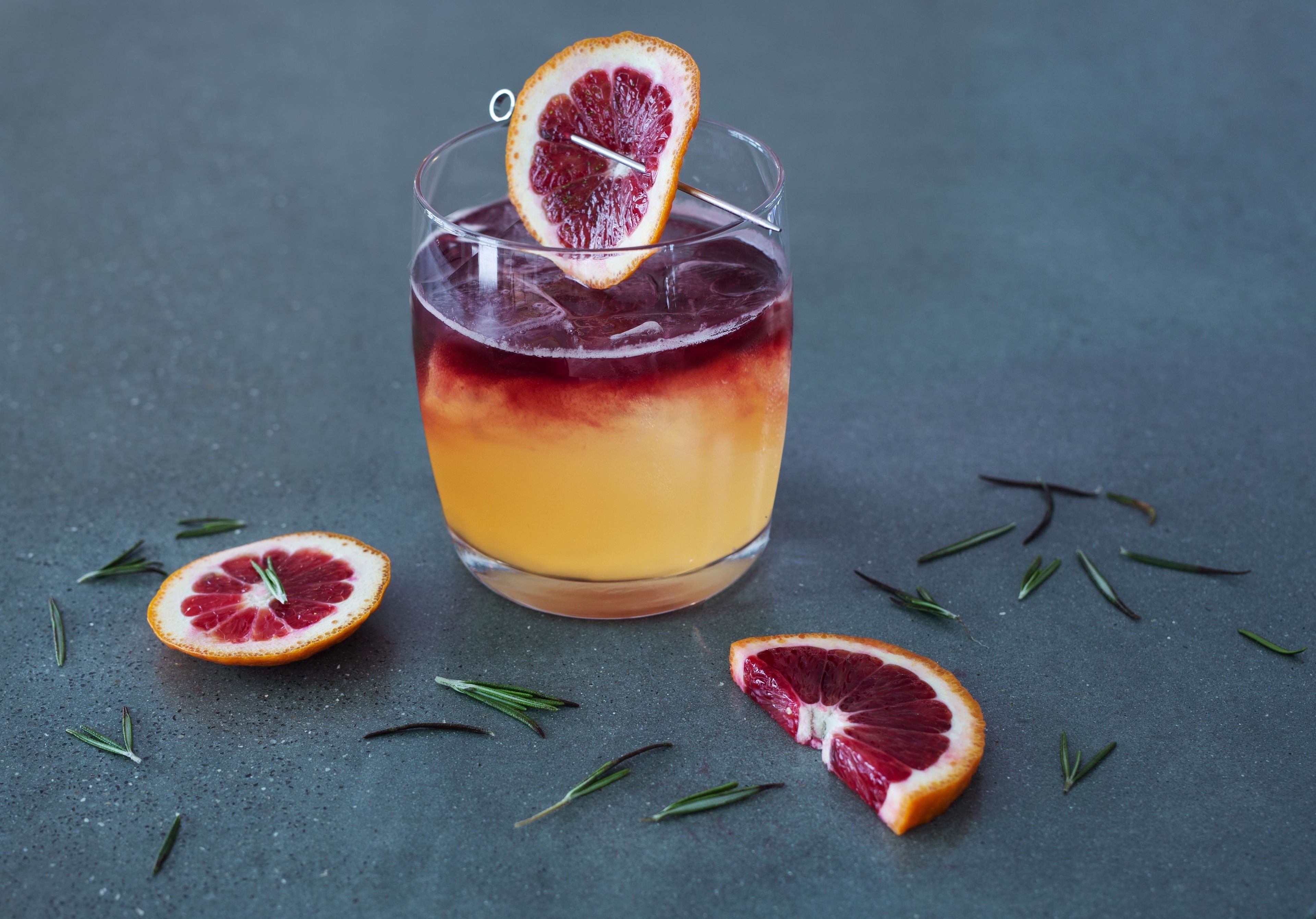 A glass of layered drink with a blood orange garnish, surrounded by scattered blood oranges and rosemary sprigs on a textured surface.