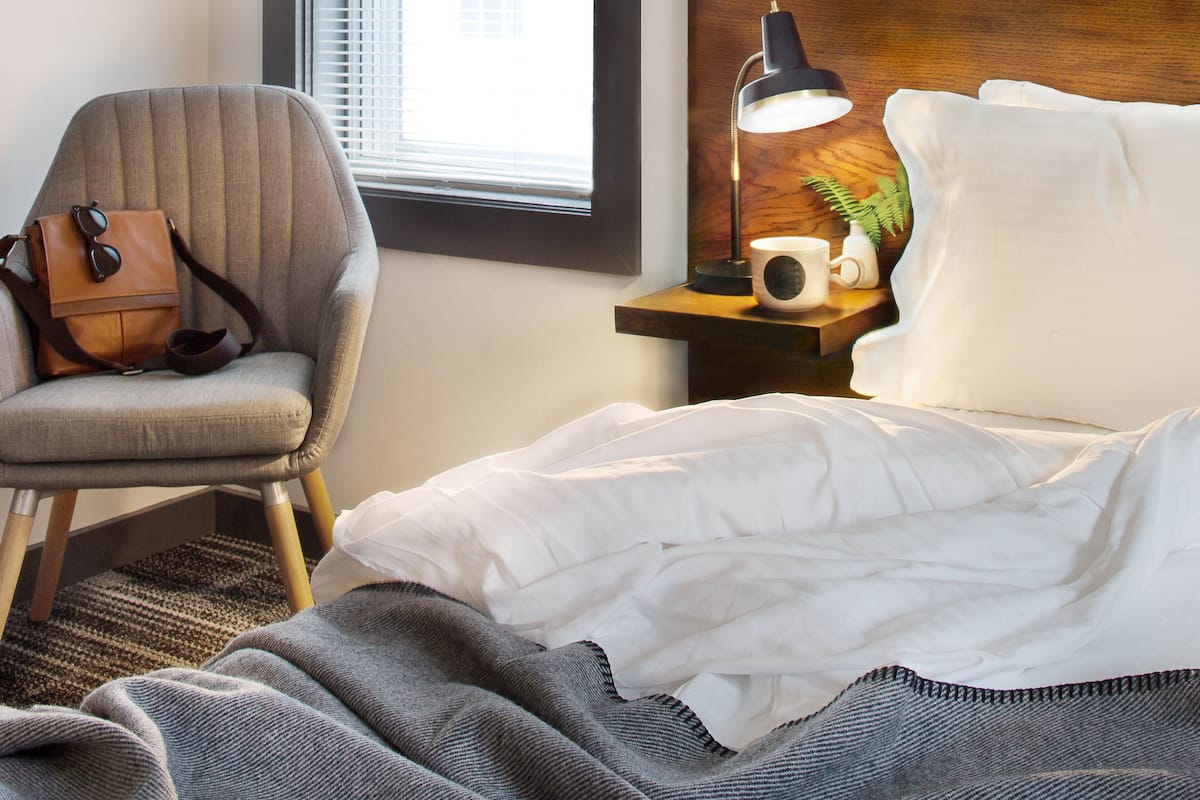 A hotel room with a bed, a chair, a nightstand with a lamp, a window, and a view of buildings outside.