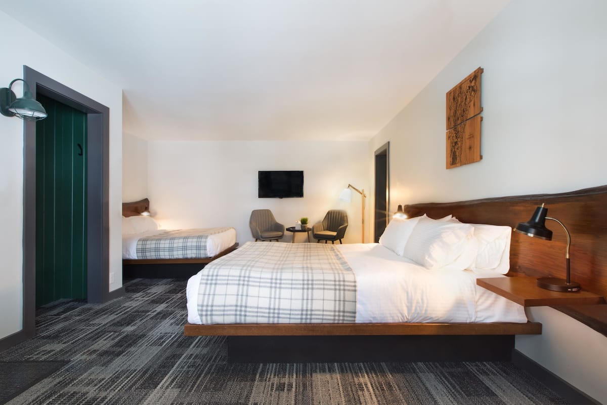 A hotel room with a large bed, a smaller bed, a desk with a lamp, two chairs, a TV, and a wooden headboard.
