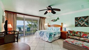 Une chambre d’hôtel avec un lit, un balcon donnant sur la mer, un ventilateur de plafond et une décoration murale sur le thème des tropiques.