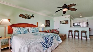 A bedroom with a bed, two bedside tables, a ceiling fan, a nightstand, a chair, a small table, and a kitchen area with a refrigerator, stove, and bar stools.