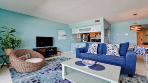 Ein Wohnzimmer mit einem blauen Sofa, einem Korbstuhl, einem Fernseher und einem durch eine offene Tür einsehbaren Küchenbereich.