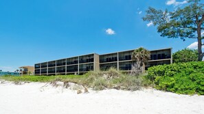 A modern building with multiple floors and large windows, situated on a sandy area with some greenery and a tree nearby.
