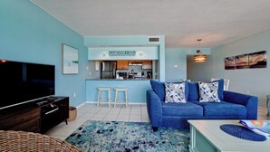 Sala de estar con un sofá azul, televisor y una zona de cocina con un letrero que dice "SANDDOLLAR".