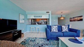 A living room with a blue sofa, a TV, and a kitchen area with a sign reading "SANDDOLLAR".