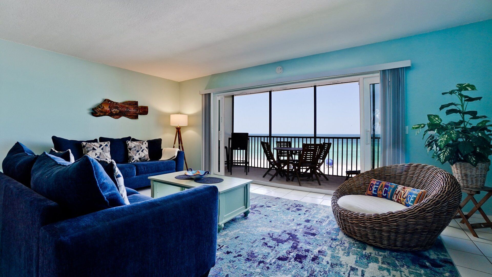 A living room with a sofa, coffee table, and a wicker chair. There is a balcony with a table and chairs, and a plant on the right.