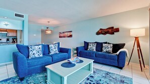 A modern living room with two blue sofas, a white coffee table, and a kitchen area with a microwave and stove.