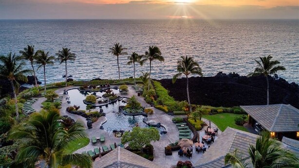 A resort with a pool area, palm trees, and a view of the ocean at sunset.
