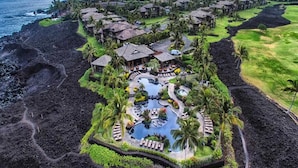 An aerial view of a resort with a central pool, multiple guest villas, and a surrounding landscape with a coastline.