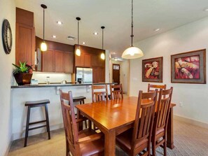 A dining area with a wooden table and chairs, a kitchen with wooden cabinets, pendant lights, and framed artwork on the wall.
