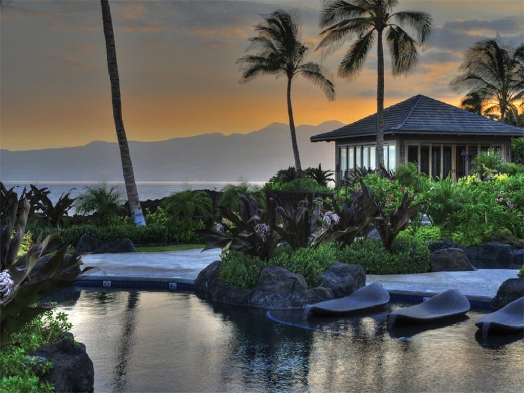 A resort with a pool, lounge chairs, palm trees, and a view of the ocean at sunset.