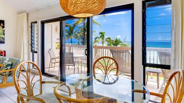 Una habitación con una mesa y sillas de vidrio, una lámpara colgante de mimbre y vistas a palmeras y a la playa a través de amplios ventanales correderas.