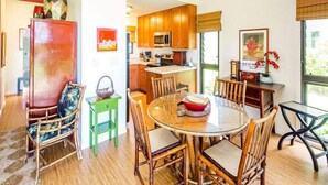 A dining area with a round glass table surrounded by wooden chairs, a kitchen with wooden cabinets and a microwave, and a view of the outdoors through a window. - Stylish Tropical Suite w/Bath+Kitchen Upgrades, WiFi, DVD, Lanai–Kaha Lani 113 (LIHUE)