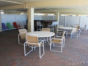 A covered outdoor seating area with round tables and metal chairs, a barbecue grill, and a white fence in the background.