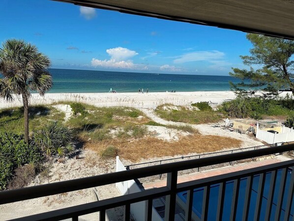 A beach view from a balcony with a clear sky, sandy beach, and a few people walking.