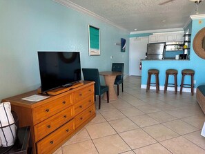 A living room with a TV, wooden dresser, teal chairs, and a small table. A kitchen with a fridge and bar stools is visible in the background.