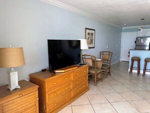 A living room with a wooden dresser, a flat-screen TV, wicker chairs, and a kitchen area with a refrigerator.