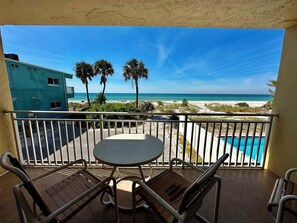 A balcony with a view of the beach, a pool, and a building in the background.