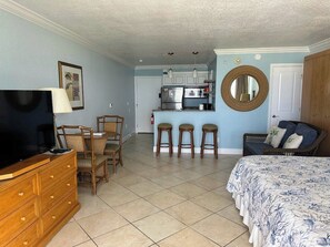 A room with a TV, wooden dresser, wicker chairs, a bed with a floral bedspread, a kitchen area with a refrigerator and microwave, and a round mirror on the wall.