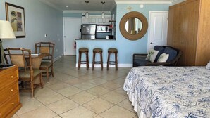 A bedroom with a bed, a dresser, a chair, a small kitchenette, and a round mirror.