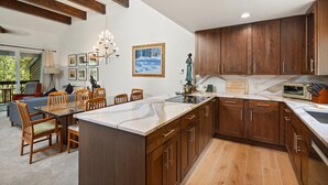 A modern kitchen with wooden cabinets, a marble countertop, and a dining area with a glass table and chairs. - Riverside Condo with Mountain and Town views and updated furnishings. (Telluride)