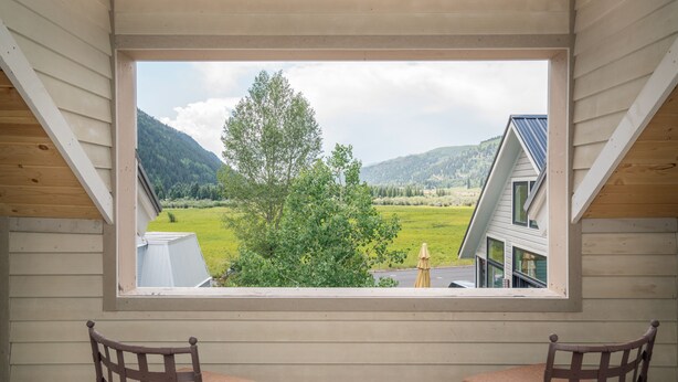 Ein Zimmer mit großem Fenster mit Blick auf eine grüne Landschaft und ein Haus. Zwei Holzstühle mit Kissen sind vorhanden.