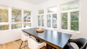 Ein Essbereich mit einem langen Tisch, weißen Stühlen und großen Fenstern mit Blick auf ein Haus und Bäume.