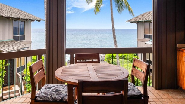 Une terrasse en bois avec une table ronde et des chaises, donnant sur une plage bordée de palmiers et offrant une vue sur un ciel dégagé.