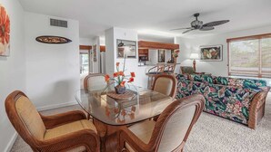 Un coin repas avec une table en verre entourée de chaises en osier, un ventilateur de plafond et, au fond, un canapé à motifs floraux.